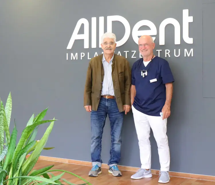 Zwei Männer stehen lächelnd vor einer Wand mit der Aufschrift AllDent Implantatzentrum.