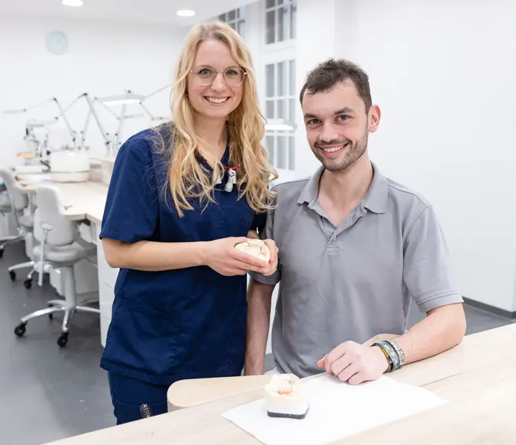 Eine Frau in blauer Arbeitskleidung steht neben einem Mann in grauem Poloshirt an einem Tisch. Beide lächeln in die Kamera, die Frau hält ein Zahnmodell in den Händen, auf dem Tisch liegt ein weiteres Modell.