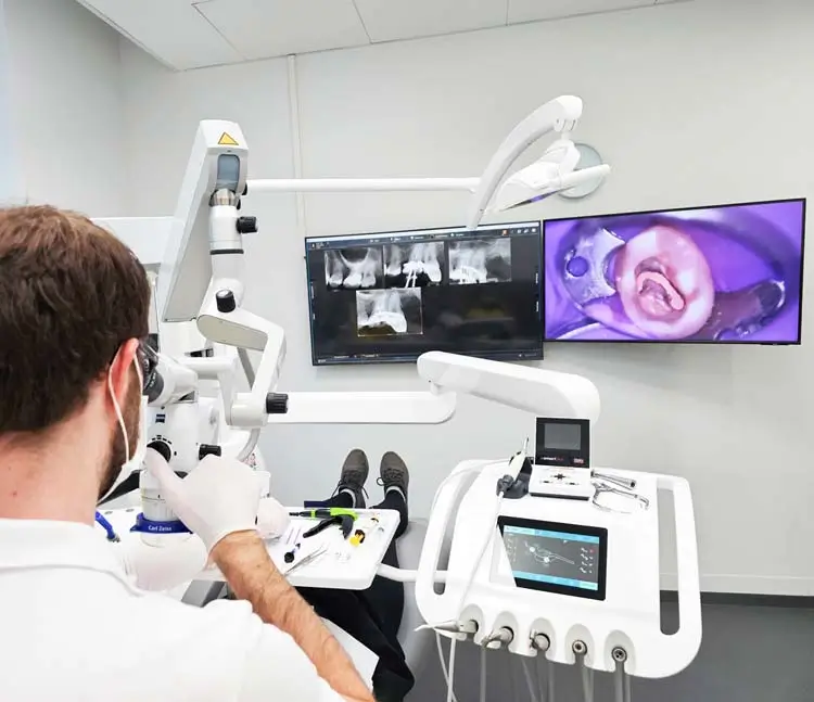 Ein Zahnarzt arbeitet mit einem Mikroskop an einem Patienten. Im Hintergrund sind Monitore mit Röntgenaufnahmen und einer Nahaufnahme eines Zahns zu sehen