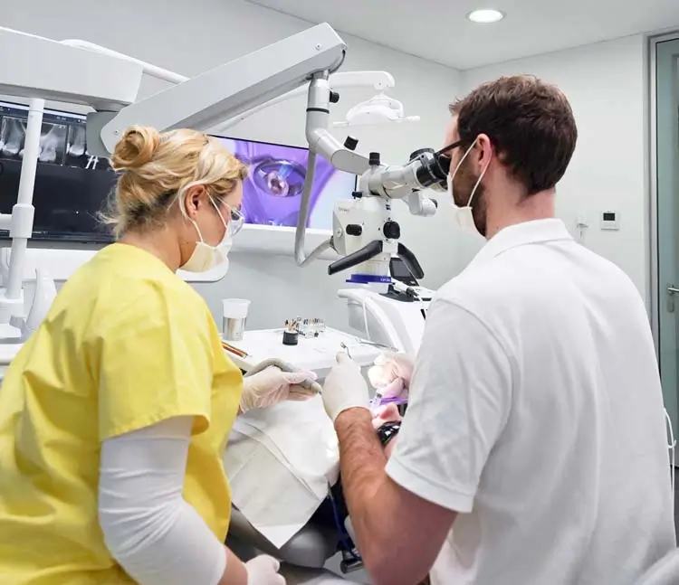Zahnarzt mit Mundschutz behandelt einen Patienten. Einer arbeitet mit einem Mikroskop, während die andere Person assistiert. Im Hintergrund sind Monitore mit Zahnaufnahmen zu sehen
