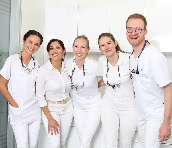 Team aus Zahnärzten steht nebeneinander im Behandlungszimmer und lächelt in die Kamera