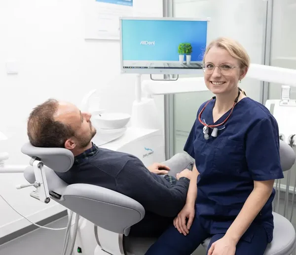 Eine Zahn&auml;rztin in blauer Arbeitskleidung sitzt neben einem Patienten der im Behandlungsstuhl aitzt und l&auml;chelt in die Kamera, im Hintergrund ist ein Monitor zu sehen