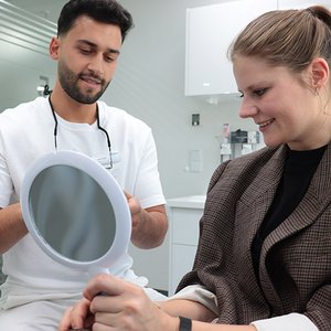 Patientin schaut in Handspiegel, Zahnarzt steht links neben ihr