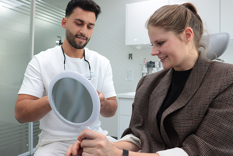 Patientin auf Behandlungsstuhl hält Handspiegel, daneben sitzt ein Zahnarzt