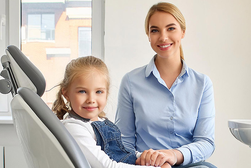 Blondes Mädchen sitzt auf Zahnarztstuhl und lächelt. Mutter sitzt auf einem Stuhl neben ihr (Bild ist KI-generiert)